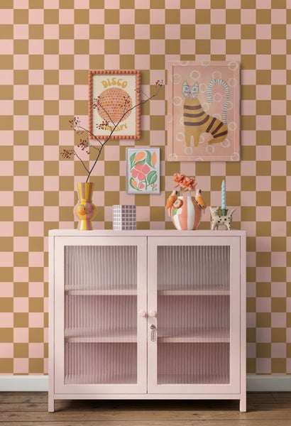 Rosa Metallschrank mit Riffelglastüren vor einer karierten Tapete mit verschiedenen Dekorationsartikeln.
