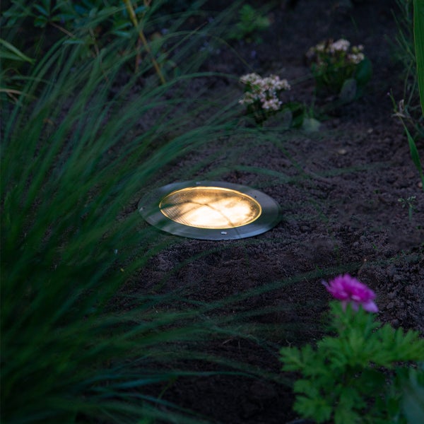 Runder Bodeneinbaustrahler aus Metall in einem Gartenbeet mit warmweißem Licht.