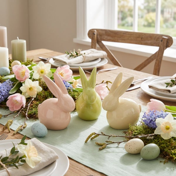 Festlich gedeckter Ostertisch mit drei Keramikosterhasen, Frühlingsblumen wie Tulpen und Narzissen, Moos und Ostereiern als Tischdekoration.