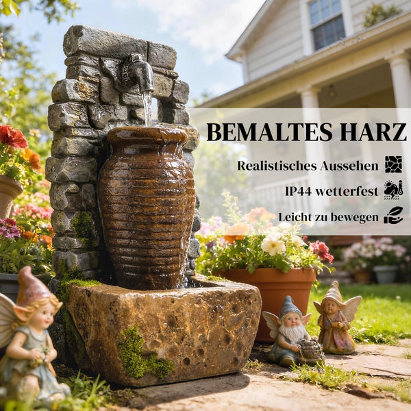 Gartenbrunnen aus wetterfestem, bemaltem Harz in Stein- und Terrakotta-Optik mit Krug und Wasserhahn in einem hellen Garten mit Blumen und Figuren.