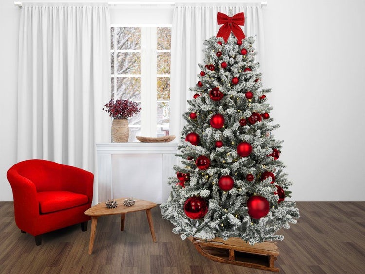 Ein künstlicher, verschneiter Weihnachtsbaum geschmückt mit Kugeln und einer Schleife, platziert auf einem Holzschlitten in einem Wohnzimmer.
