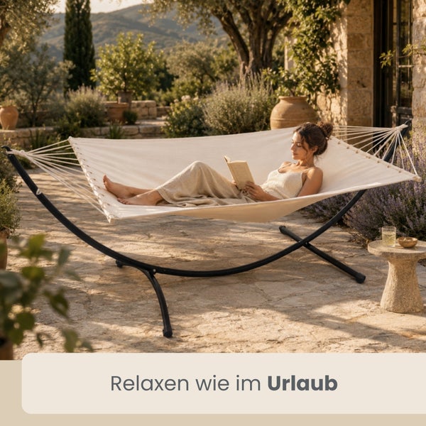 Beige Hängematte mit schwarzem Metallgestell auf einer sonnigen Steinterrasse in einem mediterranen Garten, eine Frau liegt lesend darin.