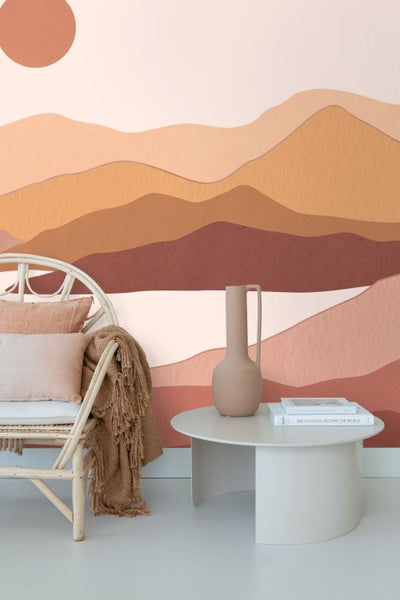 Helles Wohnzimmer mit Wandbild einer abstrakten Gebirgslandschaft und Sonne, einem Rattan-Sessel und einem Couchtisch.