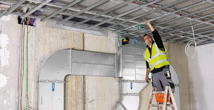 Handwerker mit Warnweste auf einer Leiter bei der Montage einer Unterkonstruktion für eine Decke mit einem Linienlaser auf einer Baustelle.