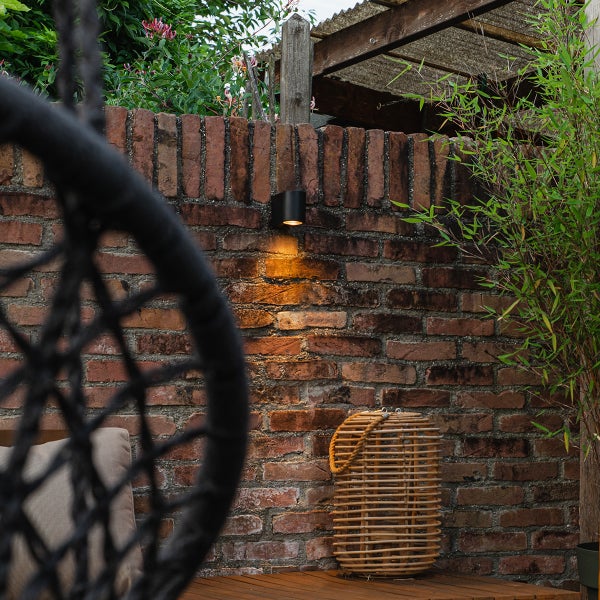 Zylindrische Außenwandleuchte an einer Backsteinmauer im Garten, nach unten gerichteter Lichtkegel.