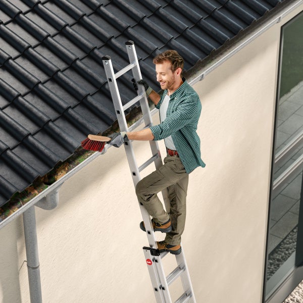 Ein Mann reinigt eine Dachrinne mit einer Bürste, während er auf einer Hailo Mehrzweckleiter an einer Hauswand steht.