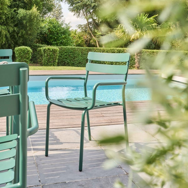 Moderner grüner Gartenstuhl aus Metall mit Armlehnen auf einer Steinterrasse an einem Pool in einem hellen Garten.