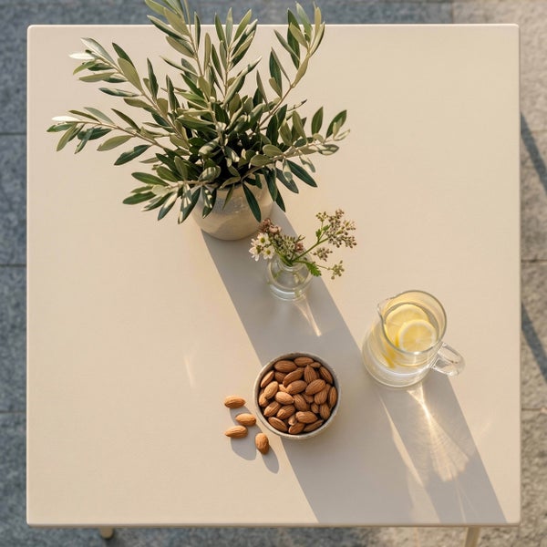 Quadratischer Gartentisch mit Olivenzweigen in einer Vase, einer Schale Mandeln und einem Glaskrug mit Zitronenwasser.