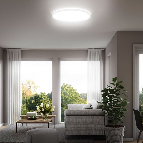 Helles, geräumiges Wohnzimmer mit runder LED-Deckenleuchte, grauem Sofa, hölzernem Couchtisch und großen Fenstern mit weißen Vorhängen.