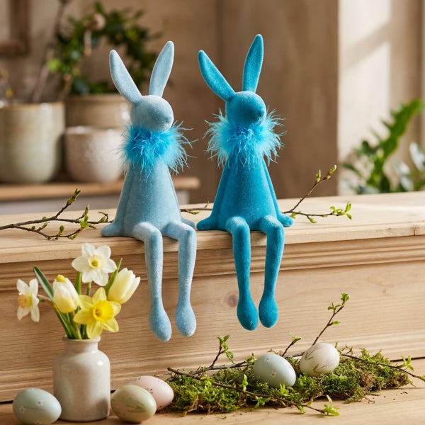 Zwei Osterhasen Figuren mit weicher Oberfläche und Federhalsband sitzen auf einer Holzbank, dekoriert mit Ostereiern und einer Vase mit Blumen.