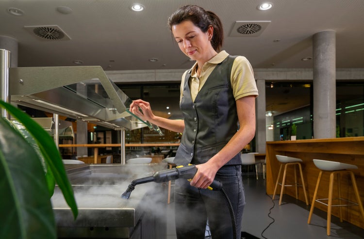 Eine Person reinigt eine Edelstahloberfläche in einem modernen Gastronomiebereich mit einem Dampfreiniger.