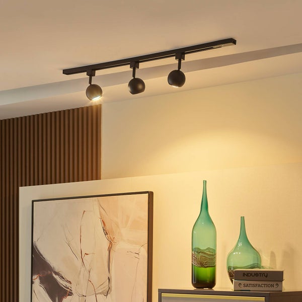 Modernes schwarzes 3-flammiges Schienensystem mit kugelförmigen Strahlern in einem hellen Raum mit abstrakter Wandkunst und grünen Glasvasen.