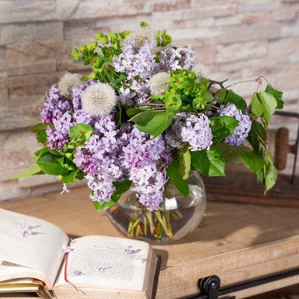 Blumenstrauß aus Flieder und Pusteblumen in einer runden Glasvase auf einem Holztisch neben einem Buch vor einer Steinwand.