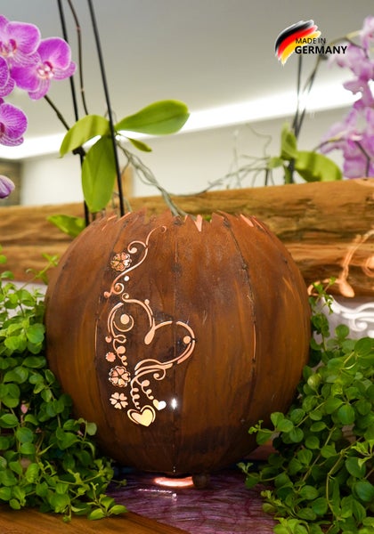 Rost-Deko-Kugel mit Herz-Motiv und Beleuchtung, platziert zwischen grünen Pflanzen und Orchideen.