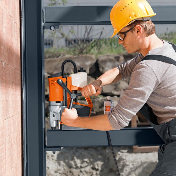 Ein Arbeiter mit Schutzhelm und Schutzbrille bedient eine Magnet-Kernbohrmaschine an einem Metallträger im Außenbereich.