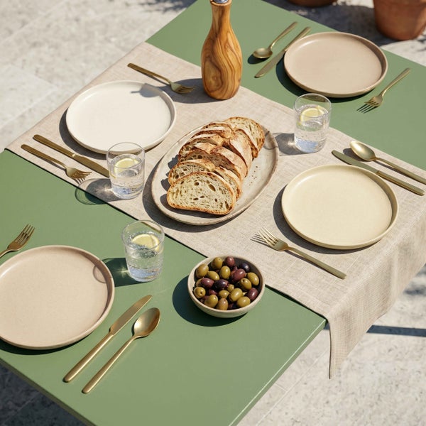 Gedeckter Gartentisch mit beigen Tellern, goldenem Besteck, Brotplatte, Olivenschale und Wassergläsern auf grünem Tisch mit hellem Leinenläufer.