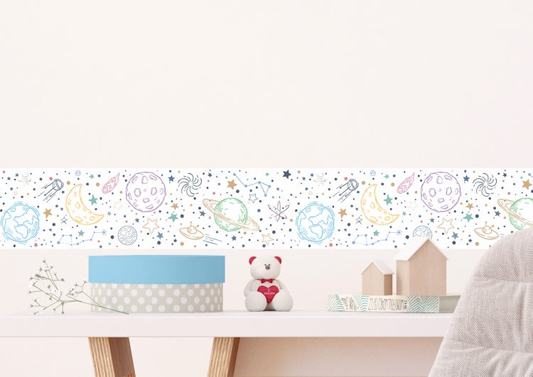 Selbstklebende Tapetenbordüre mit Weltraum-Motiven wie Planeten und Sternen an einer Wand über einem Tisch in einem hellen Kinderzimmer.