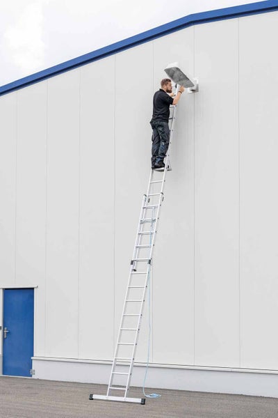 Eine Person steht auf einer Aluminium-Mehrzweckleiter und montiert eine Aussenleuchte an einer Fassade.