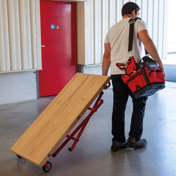 Eine Person zieht eine mobile Werkbank mit Holzplatte und rotem Metallgestell, während sie eine rote Werkzeugtasche über der Schulter trägt.