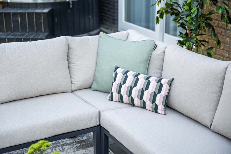 L-förmiges Garten-Lounge-Sofa mit hellgrauen Polstern, einem salbeigrünen Dekokissen und einem gemusterten Kissen auf einer Terrasse.