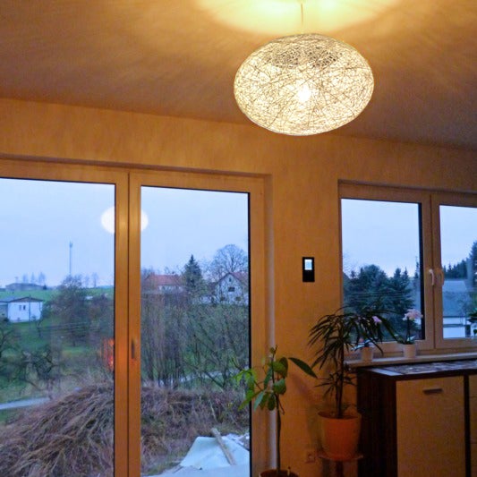 Moderne Pendelleuchte mit rundem, fein geflochtenem Schirm in einem hell erleuchteten Wohnzimmer mit großen Fensterfronten.