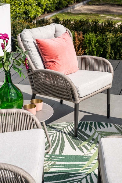 Moderner Garten-Loungesessel mit Seilgeflecht, hellgrauen Polstern und einem Dekokissen auf einer Terrasse mit Palmenblatt-Teppich.