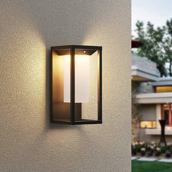 Moderne Außenwandleuchte mit schwarzem Metallrahmen und weißem Diffusor, montiert an einer strukturierten Hauswand, erzeugt warmes Licht.