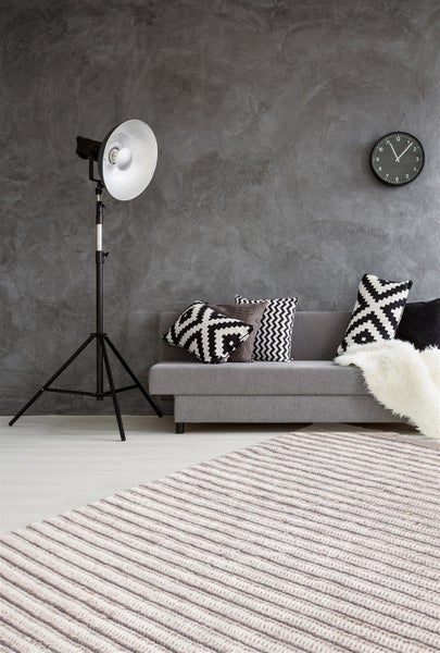 Modernes Wohnzimmer mit grauem Sofa, strukturierten Kissen, Stehlampe im Studio Stil, Wanduhr und gestricktem Teppich vor einer strukturierten Wand.