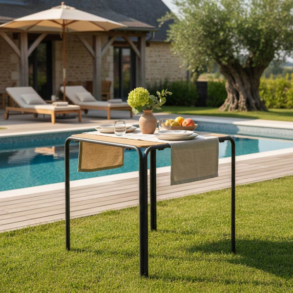 Kleiner quadratischer Gartentisch mit schwarzem Metallgestell und Holzplatte, gedeckt für eine Mahlzeit neben einem Pool auf einer hellen Terrasse.
