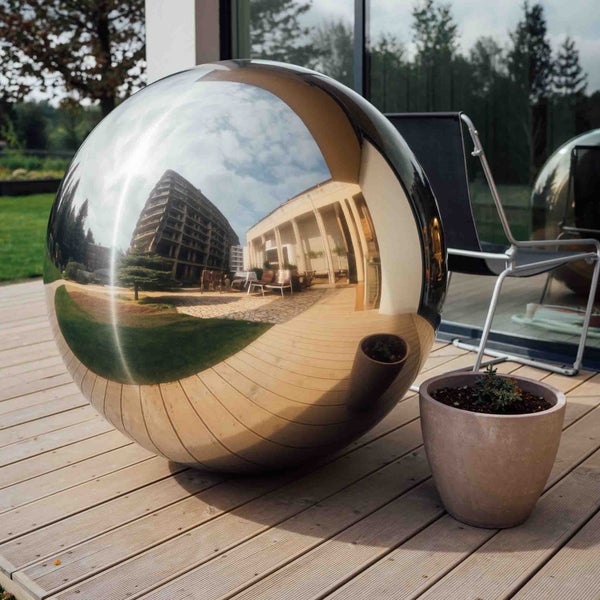 Spiegelnde Gartenkugel aus Edelstahl auf einer hellen Holzterrasse in einem modern gestalteten Außenbereich mit Pflanzkübel.