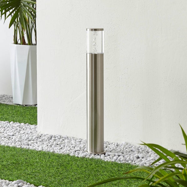 Moderne Wegeleuchte aus Edelstahl mit transparentem Aufsatz mit Luftblasen auf Kieselsteinen neben Kunstrasen vor einer Wand.