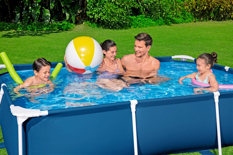 Familie spielt in einem rechteckigen blauen Rahmenpool auf einer Rasenfläche mit einem Wasserball und Poolnudeln.