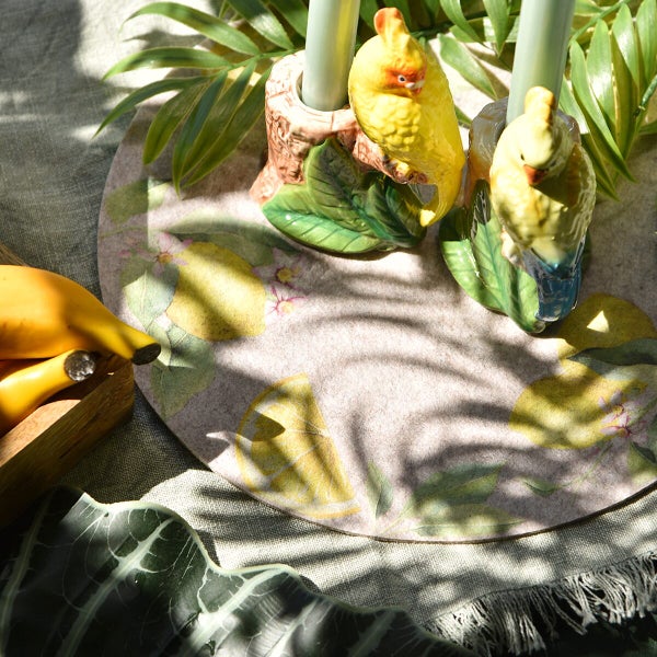 Zwei Kerzenhalter in Papageienform auf einem runden Tischset mit Zitronenmotiv, dekoriert mit grünen Pflanzen und Früchten.