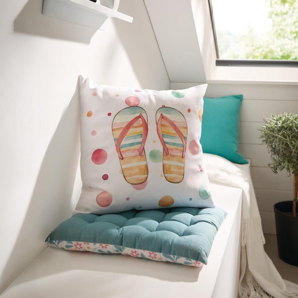Zierkissen mit Flip-Flop-Motiv und bunten Punkten auf einem blaugrünen Sitzkissen in einem hellen Raum am Fenster.