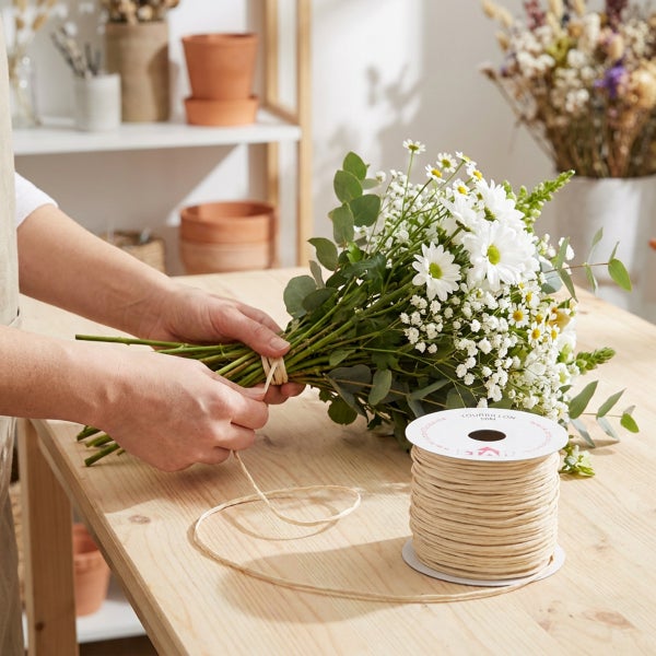 Eine Person bindet einen Blumenstrauß aus Gänseblümchen und Schleierkraut mit einer Naturkordel auf einem Holztisch.