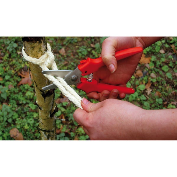 Hände schneiden mit einer Mehrzweckschere ein dickes Naturseil an einem jungen Baumstamm im Garten.