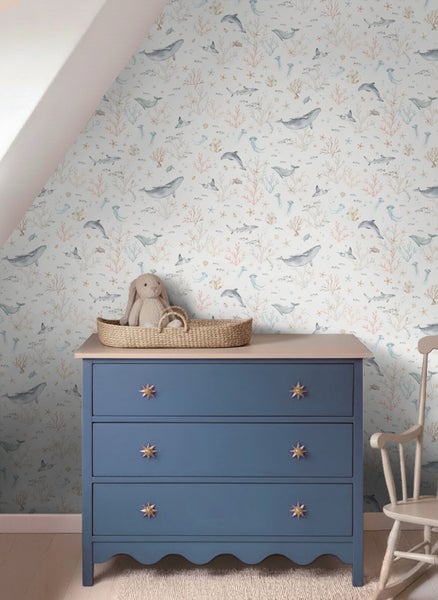Kinderzimmer mit maritimer Tapete mit Walen und Delfinen, blauer Kommode und Weidenkorb in einem hellen, aufgeräumten Raum.