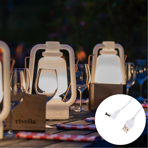 USB-Ladekabel mit DC-Anschluss fuer kabellose Aussenleuchten, im Hintergrund beleuchtete Tischlaternen auf einem Holztisch bei Nacht.
