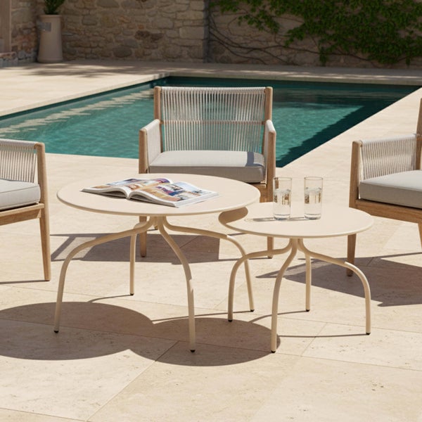 Zwei runde Beistelltische für den Außenbereich in Beige auf einer hellen Terrasse neben einem Swimmingpool und Gartenstühlen aus Holz.