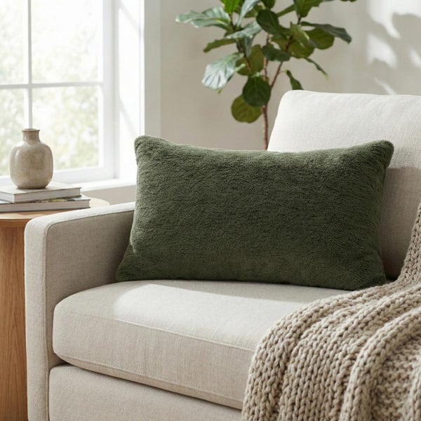 Grünes, rechteckiges Dekokissen mit weicher Textur auf einem hellen Sessel neben einem Fenster und einer Zimmerpflanze.