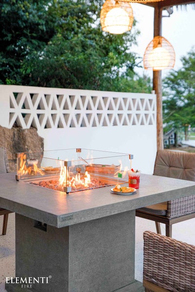 Moderner quadratischer Feuertisch aus grauem Steinimitat mit Glas-Windschutz und offener Flamme auf einer Terrasse mit Gartenmöbeln.
