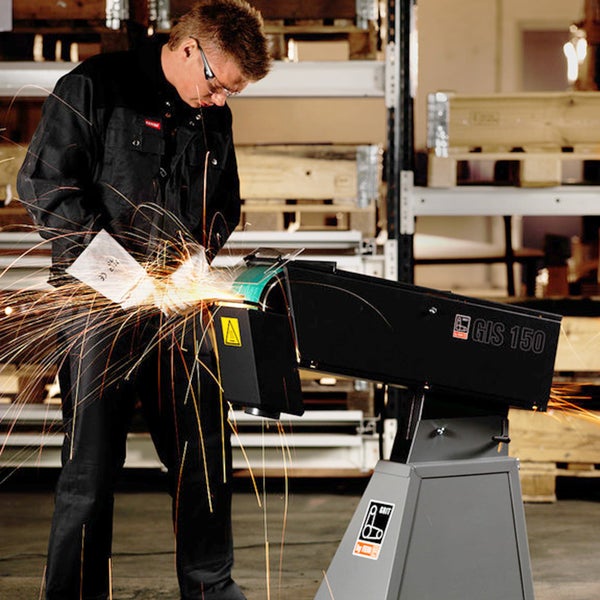 Ein Handwerker in Schutzkleidung arbeitet an einer stationären Bandschleifmaschine FEIN GIS 150 mit Funkenflug beim Metallschleifen in einer Werkstatt.