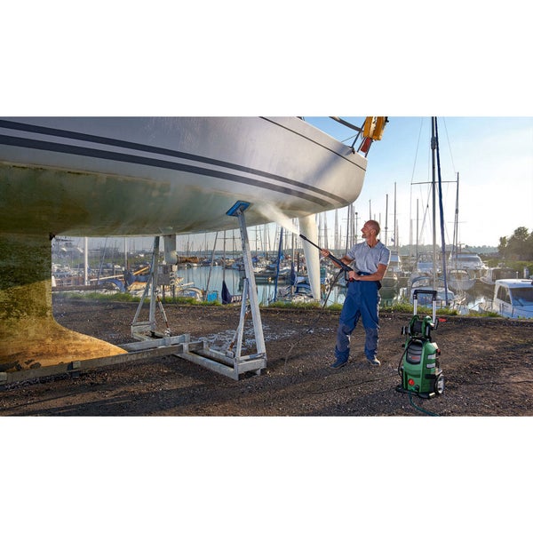 Ein Mann reinigt den Rumpf eines Segelboots mit einem Hochdruckreiniger in einem Hafenbecken.