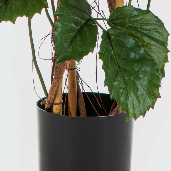 Künstliche Pflanze im schwarzen Topf mit gezackten grünen Blättern und Holzstielen.