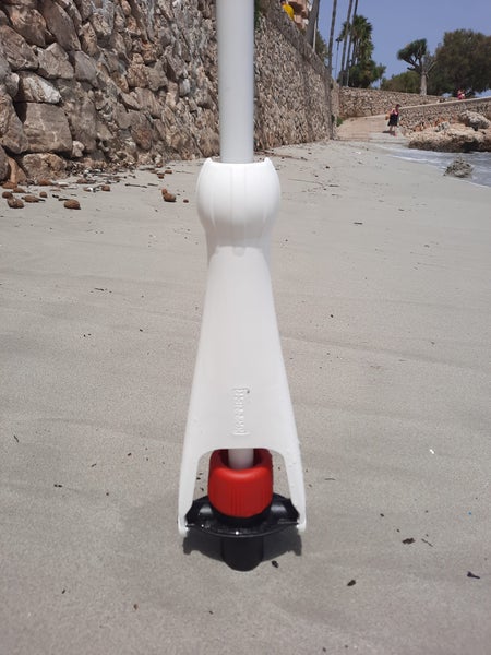 Weißer Krinner Sonnenschirmständer aus Kunststoff mit rotem Drehknopf, fest im Sandboden am Strand verankert.
