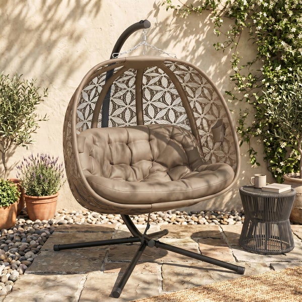 Hängesessel mit Metallgestell und Geflechtkorb inklusive weichem Sitzpolster auf einer Terrasse mit Steinboden und Gartenpflanzen.