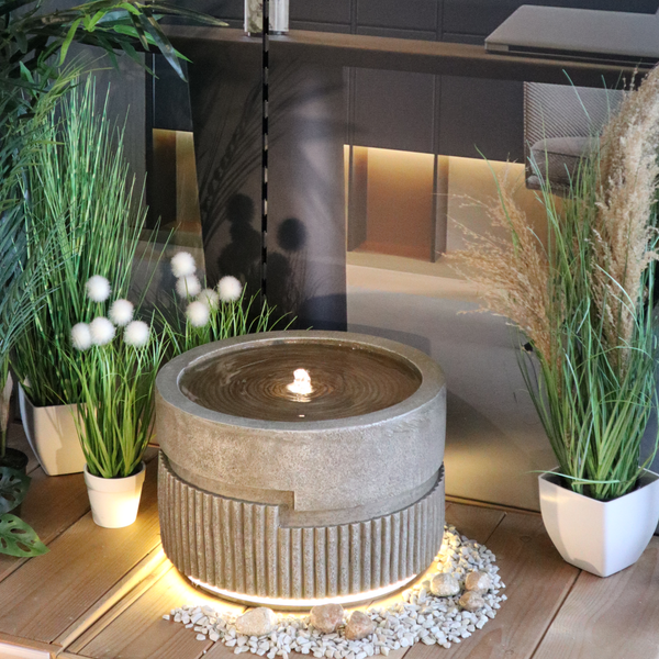 Moderner Zimmerbrunnen in Betonoptik mit integrierter Beleuchtung am Sockel, umgeben von Zierpflanzen und Kieselsteinen auf einem Holzdeck.