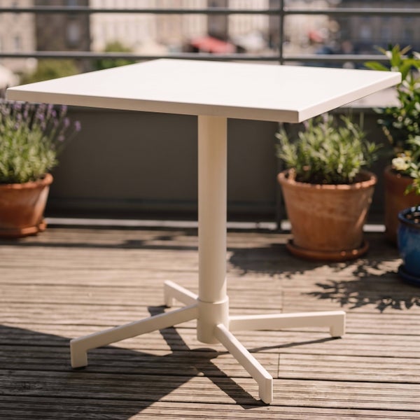 Quadratischer, cremeweißer Bistrotisch auf einer Holzterrasse mit Pflanzen im Hintergrund.