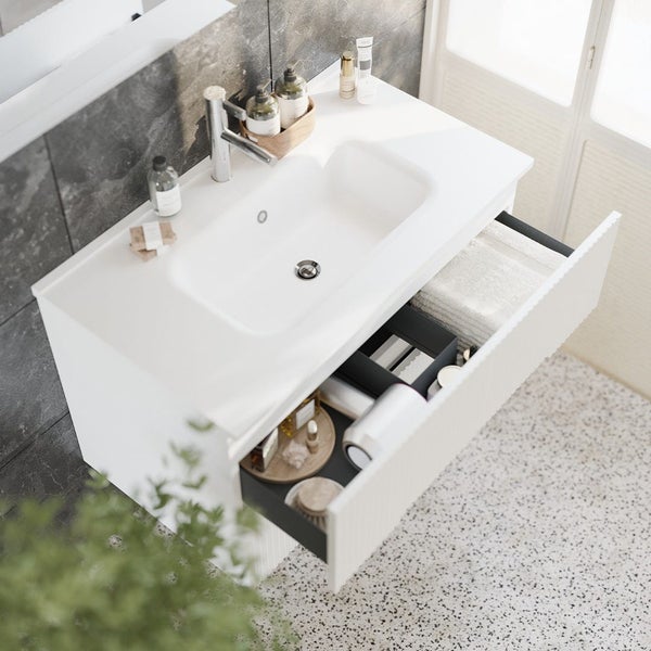 Moderner Waschtisch mit weißem Waschbecken, Armatur und offener Schublade mit geordnetem Inhalt vor einer dunklen Steinoptikwand im Badezimmer.