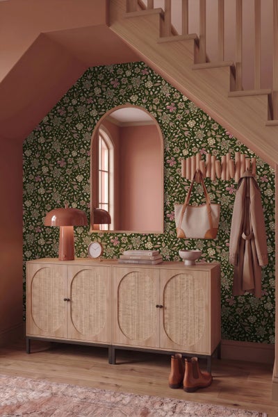 Organisierter Eingangsbereich unter einer Treppe mit dunkler Blumentapete, Sideboard aus Holz mit Rattantüren und großem Bogen-Spiegel.